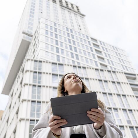 Blonde woman holding iPad looking up towards white apartment building.