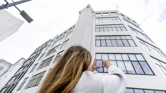 Une femme lève les mains vers un immeuble blanc de hauteur moyenne avec de nombreuses fenêtres.