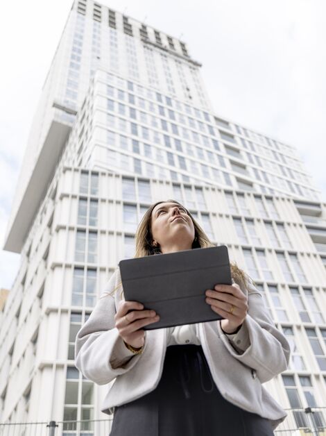 Une blonde tenant un iPad qui regarde vers un immeuble blanc.