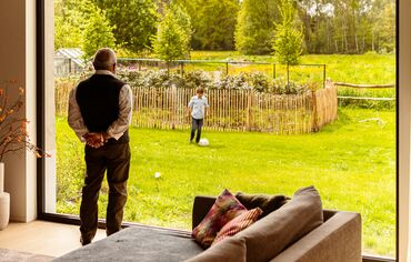 Elder person watching his grandson play in the garden from behind window wall.